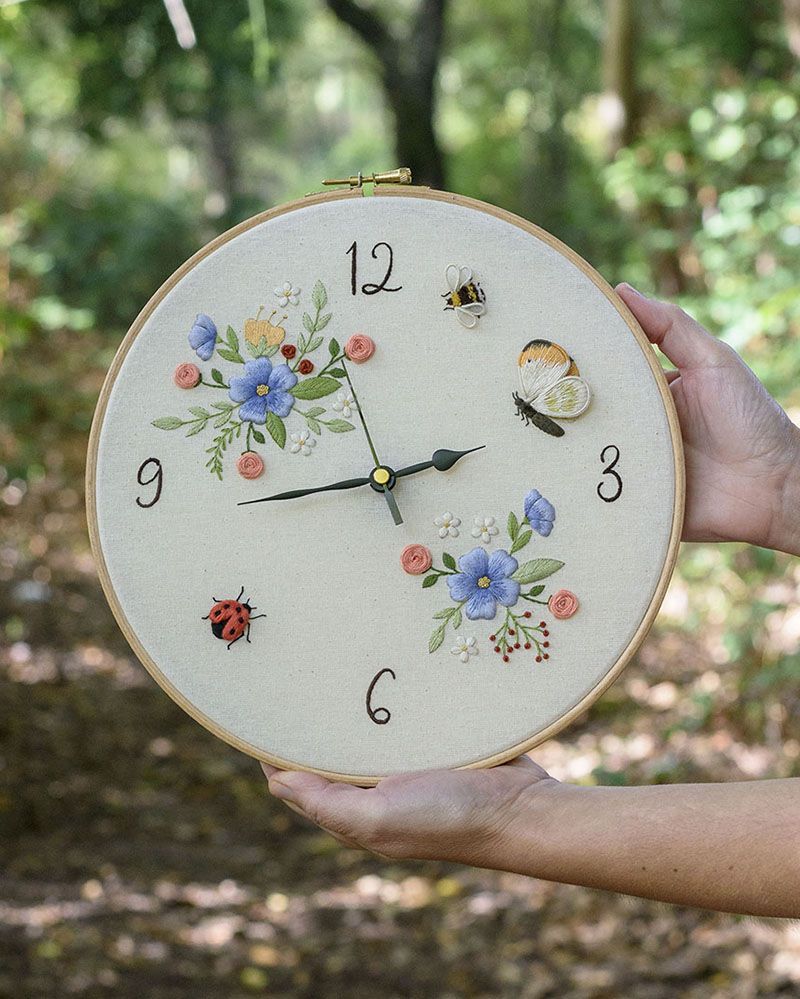 Manos de artesana sosteniendo un reloj de bastidor bordado con flores, mariposa, abeja y mariquita sobre fondo de bosque - Arorua